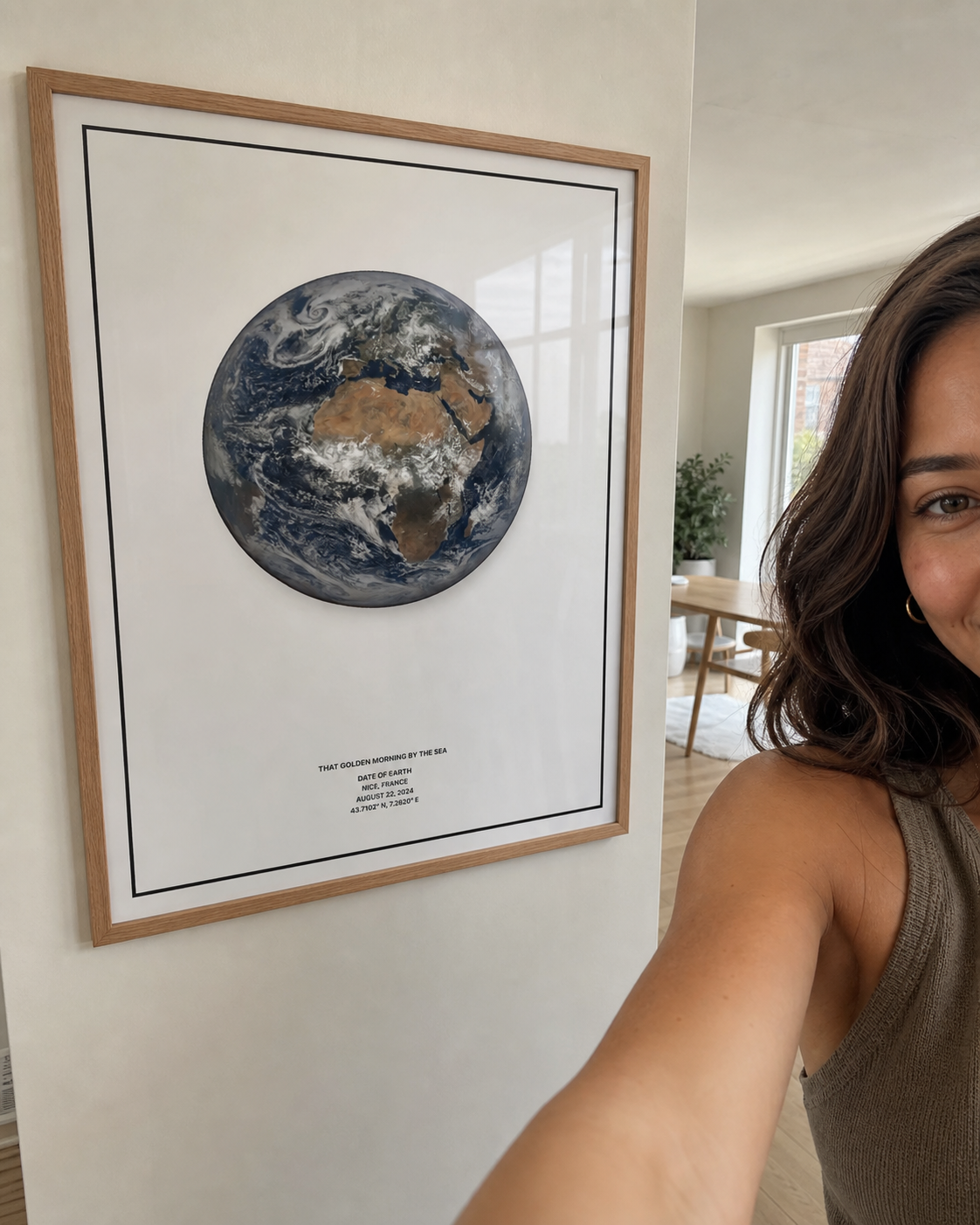 Woman taking a selfie beside a framed white Date of Earth print in a bright home.